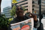 Man holds up "street sense" newspaper on city street.