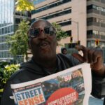 Man holds up "street sense" newspaper on city street.